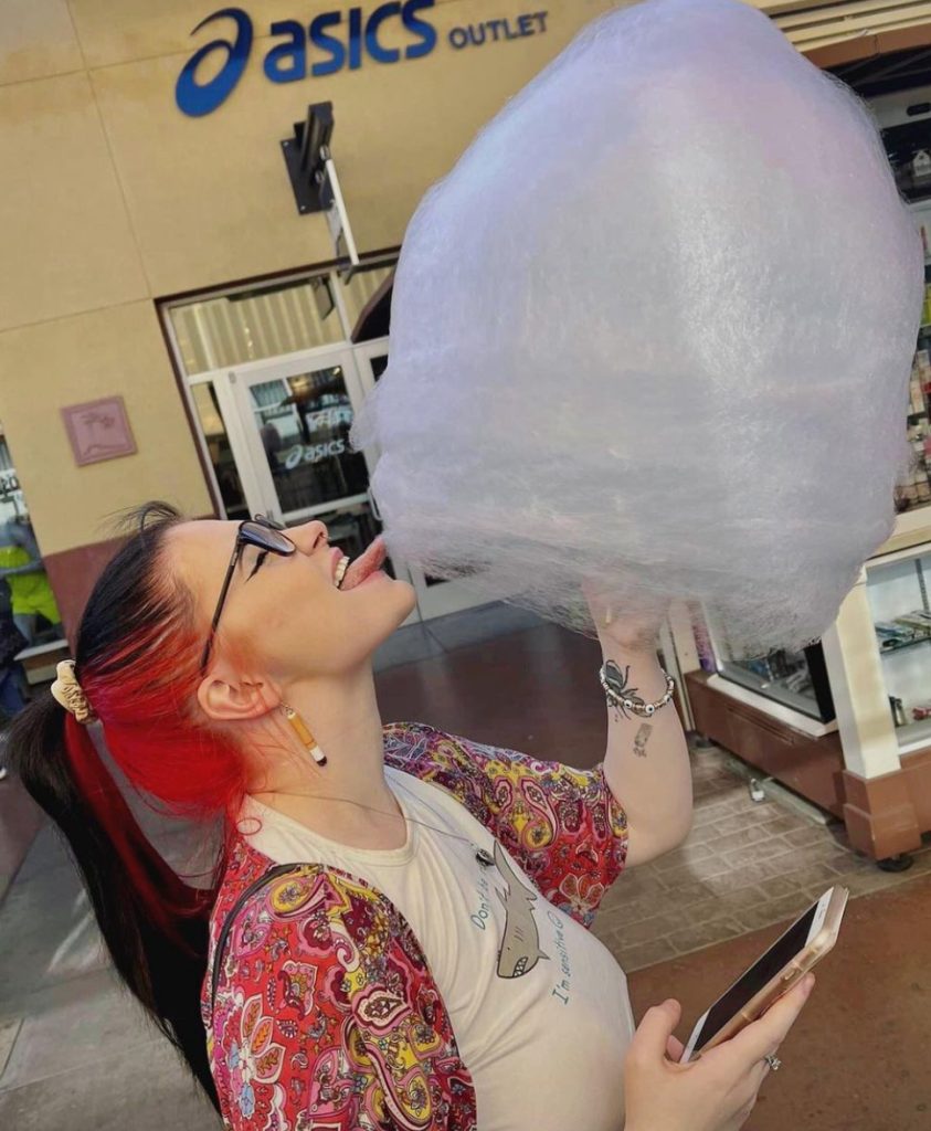 A photo of me stood at a side angel holding a giant cotton candy on a stick. My hair is red and black and is in a ponytail and I am wearing glasses